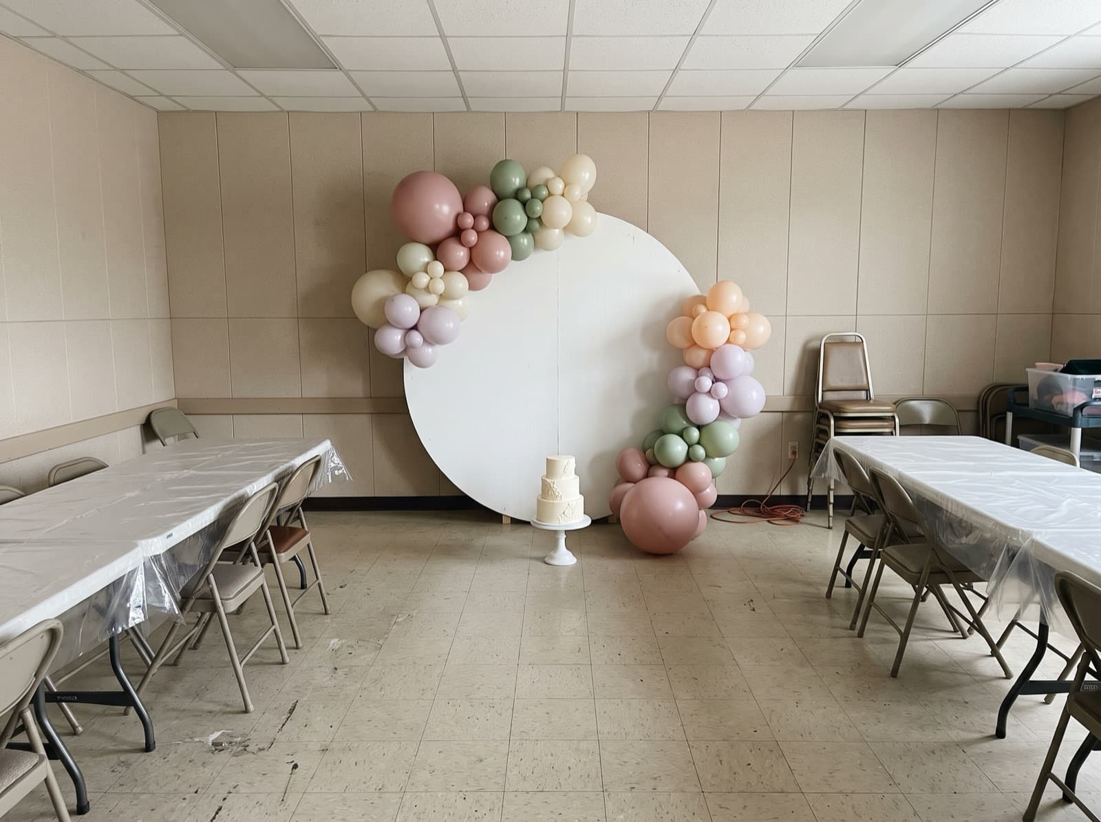 Round balloon backdrop with a pastel organic garland and a cake stand at a first birthday party in a fire hall